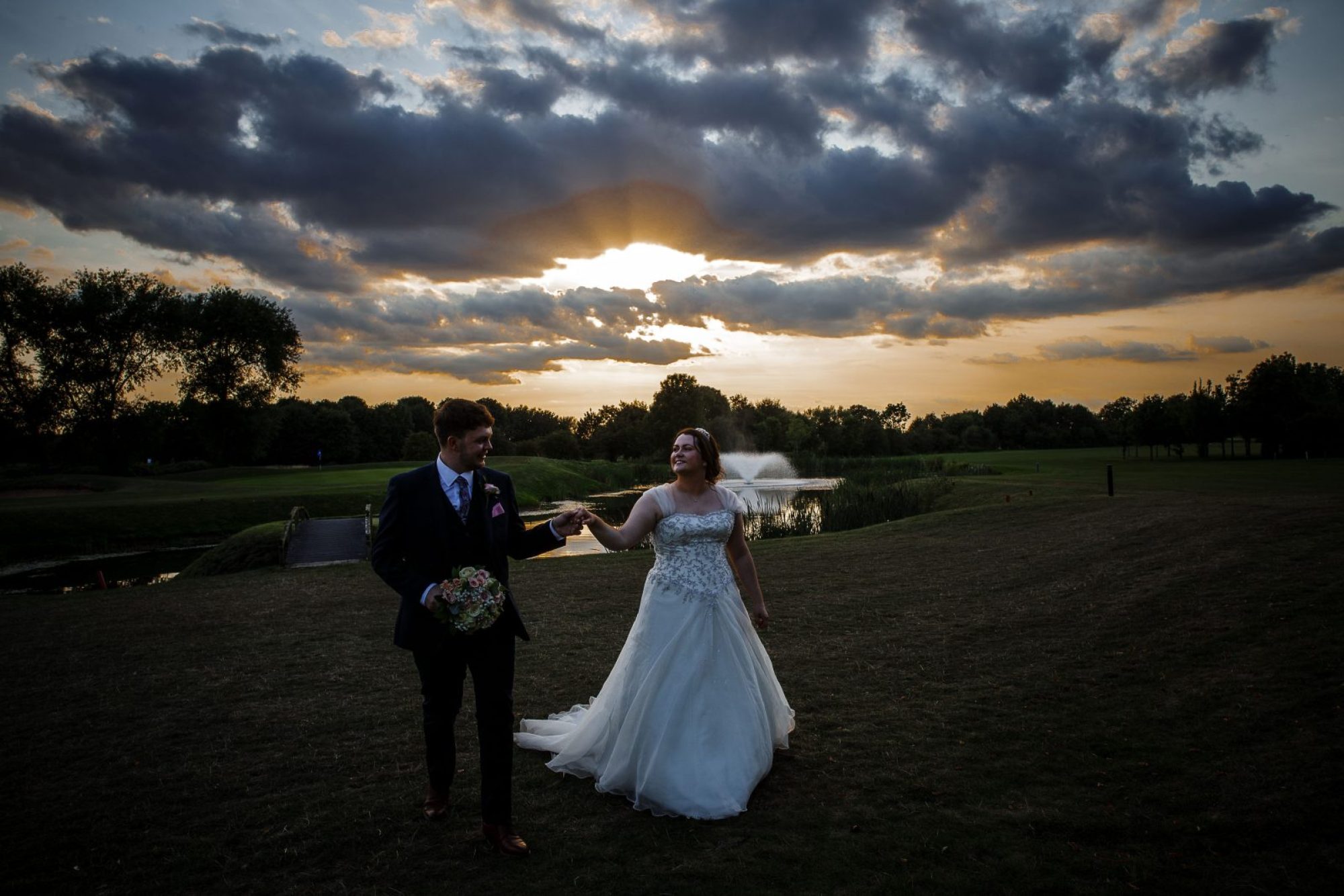 Happy couple at their wedding in Nottingham holding hands walking across the green landscape at sunset